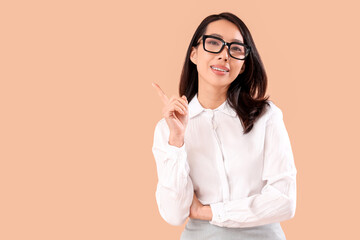 Young Asian businesswoman in eyeglasses pointing at something on beige background - obrazy, fototapety, plakaty