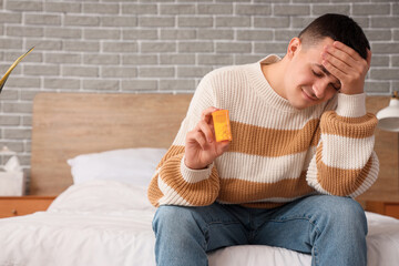 Young man with headache holding pill bottle in bedroom