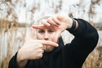 A young man looking through finger frame on natural background near river in reeds.