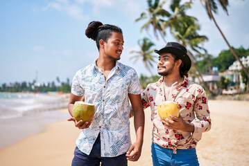 Gay couple enjoy fresh coconut water, stroll on sunny tropical beach. Men in casual summer shirts, hats, bond on romantic getaway. LGBT travel, relaxed vacation vibes, sea, palms in background.