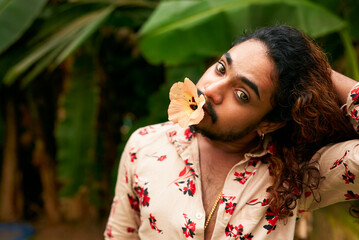 South Asian man with expressive eyes, flower in mouth, models in tropical setting. Flamboyant gay...