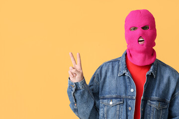 Handsome young man in balaclava showing victory gesture on yellow background
