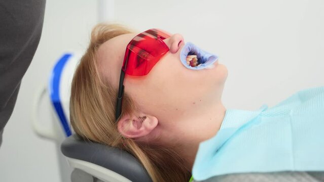 Dentist's hands puts protective red glasses on female patient's face before UV teeth whitening. Blonde woman prepares for bleaching teeth in the modern dental clinic. Cosmetic dentistry. Close-up.
