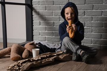 Homeless little boy eating bread near grey brick wall