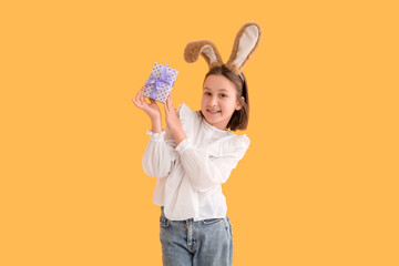Happy little girl in bunny ears with Easter gift box on yellow background