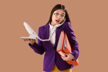 Shocked Asian businesswoman with folders talking by phone on brown background