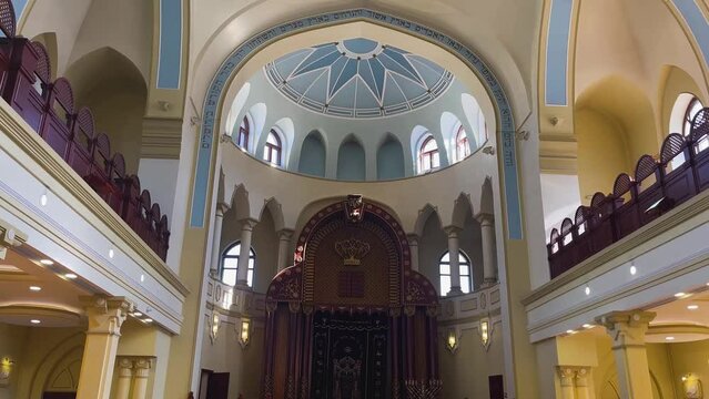 The synagogue building is from the inside. A two-storey hall with a domed ceiling. The sun's rays enter through many windows. A religious building for Jewish prayers.