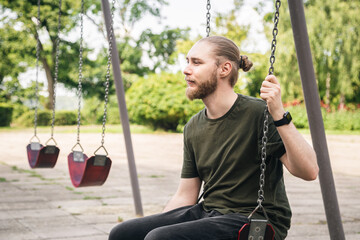 A young man with a beard is resting in the park, swinging on a swing, a man on the playground.