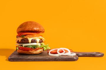 Board with tasty burger, onion rings and tomatoes on orange background