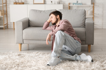 Tired young man sitting near sofa at home - obrazy, fototapety, plakaty