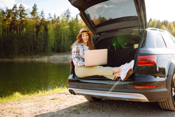 Young woman sits in the trunk of a car and works on a laptop during her journey through the highlands. Work and travel.