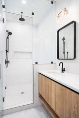 A bathroom detail with a floating wood cabinet, black faucets and mirror, white marble countertop, and a white marble tiled shower with niche.