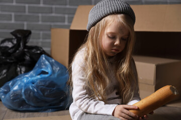 Homeless little girl eating bread near trash against grey brick wall