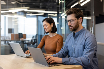 Focused man and woman using laptops at a wooden table in a bright, contemporary workspace, embodying professionalism and teamwork.