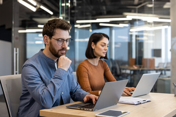 Two focused professionals working together at a desk with laptops in a contemporary office setting,...