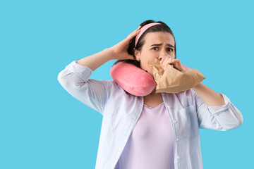 Beautiful young stressed woman with travel pillow and paper bag having panic attack on blue background. Stress Awareness Month - obrazy, fototapety, plakaty