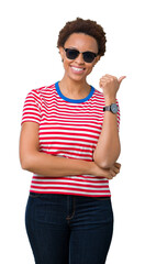 Beautiful young african american woman wearing sunglasses over isolated background smiling with happy face looking and pointing to the side with thumb up.