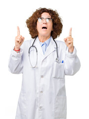 Middle ager senior doctor woman over isolated background amazed and surprised looking up and pointing with fingers and raised arms.