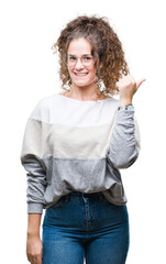 Beautiful brunette curly hair young girl wearing glasses over isolated background smiling with happy face looking and pointing to the side with thumb up.