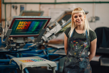 Portrait of happy print shop worker standing near screen printing press