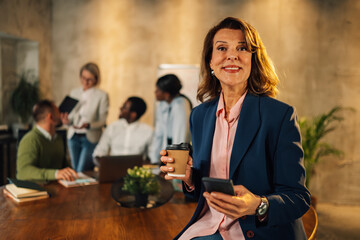 Smiling mature businesswoman with coffee and phone in hands on a meeting