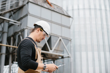 A worker in a protective helmet near grain silos looks at documents. © dsheremeta