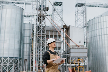 A young engineer in a hard hat with a project in his hands checks the status of the silo...