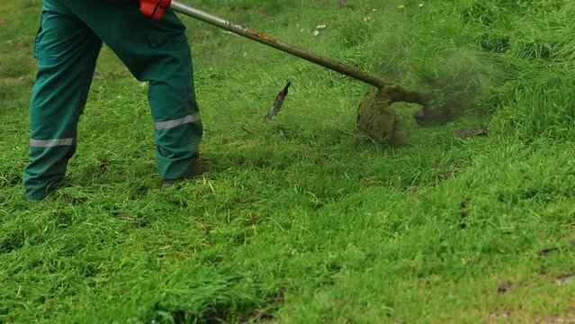 A groundskeeper in a reflective vest and protective gear is operating a weed whacker to maintain and trim the grass in a park area