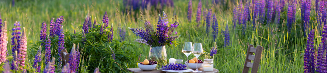 Romantic table decor for a loving couple on the blooming meadow with purple lupines. Two glasses of...