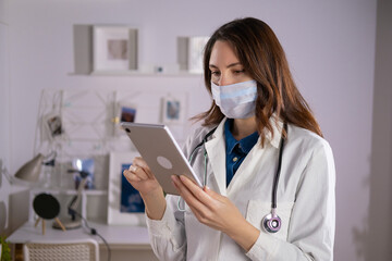 A female doctor in a mask and white uniform uses a tablet in her work. Technologies