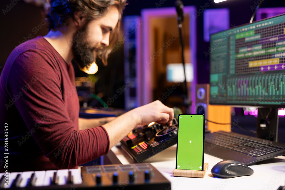Poster Music producer uses controls and knobs on mixing console in home studio, having a smartphone with greenscreen display. Artist learning to edit tunes and add sound effects with daw software.
