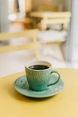 Cup of black coffee in turquoise ceramic cup on a yellow table in a cafe.