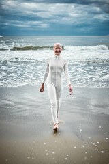 Smiling hairless girl with alopecia in white futuristic suit comes out of cold restless sea on...