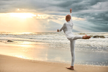 Young hairless ballerina with alopecia in white futuristic suit dancing on seashore at sunset sea,...