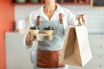 Closeup of waitress holding and serving paper cup of coffee and takeaway food in paper bag to customer in cafe, cropped shot