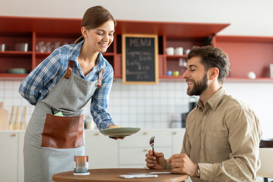 Cheerful young waitress serving some dessert to happy hungry male customer in cafe