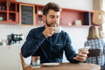 Bearded young man drinking coffee in cafe and using mobile phone, scrolling social media feed or chatting with girlfriend