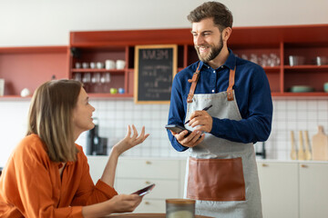 Handsome smiling waiter using application on digital tablet, taking order of young female client in pastry shop of cafe