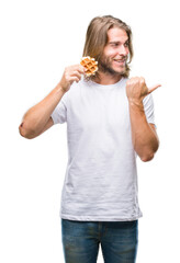 Young handsome man with long hair over isolated background eating waffle pointing and showing with thumb up to the side with happy face smiling