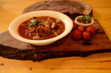 On a wooden board, a small white bowl, chicken gizzards in sauce, cherry tomatoes, basil and a container with pepper sauce and mint sprigs.