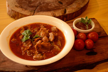 On a wooden board, a small white bowl, chicken gizzards in sauce, cherry tomatoes, basil and a container with pepper sauce and mint sprigs.