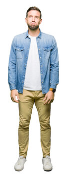 Handsome man with blue eyes and beard wearing denim jacket puffing cheeks with funny face. Mouth inflated with air, crazy expression.