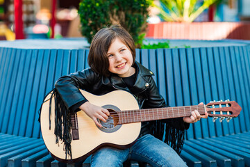 A beautiful little girl, in rock style,in a leather jacket and jeans,sits and plays an acoustic...