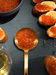 Salmon caviar in a golden spoon and a dark bowl on a dark gray stone background. Red caviar sandwiches with white wine. Gourmet food up close. Seafood and delicatessen.