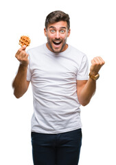 Young handsome man eating a sweet waffle over isolated background screaming proud and celebrating victory and success very excited, cheering emotion