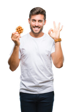 Young handsome man eating a sweet waffle over isolated background doing ok sign with fingers, excellent symbol