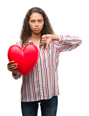 Young hispanic woman in love holding red heart with angry face, negative sign showing dislike with thumbs down, rejection concept