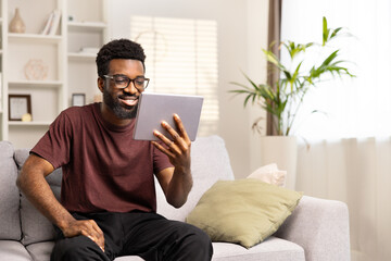 Cheerful Young Man Enjoying Tablet On Cozy Sofa At Home. Positive African American Smiling, Casual,...