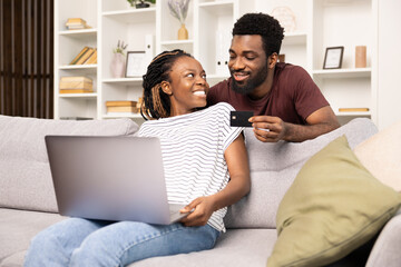 A cheerful couple is seen shopping online from their couch at home, using a laptop and a credit card, sharing a joyful moment.