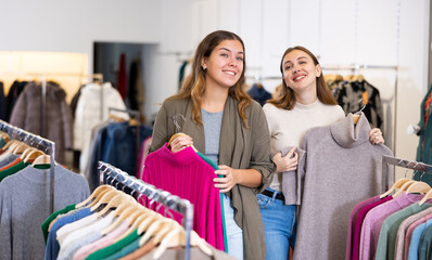 Cheerful females holding warm jumpers and sweaters on hangers while spending time together in a...